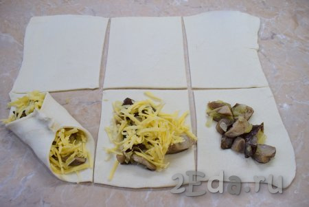 На каждый квадратик теста выложите грибную начинку и посыпьте натёртым сыром. Два противоположных уголка каждого квадрата теста соедините в центре, как на фото, формируя слойку с грибами и сыром. Так сформируйте все слойки.