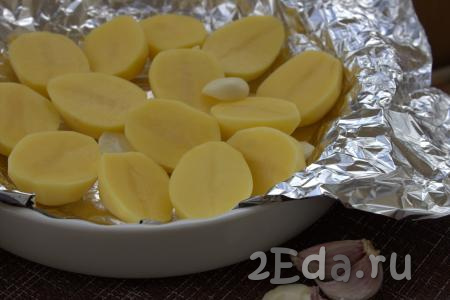 Чеснок очистить и разрезать на две части. В жаропрочную форму, застеленную фольгой, выложить в один слой половинки картофеля, поверх картошки разместить нарезанный чеснок.
