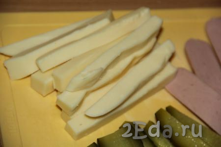 Сыр нарезать на брусочки такой же длины, как нарезаны сосиски и огурцы.