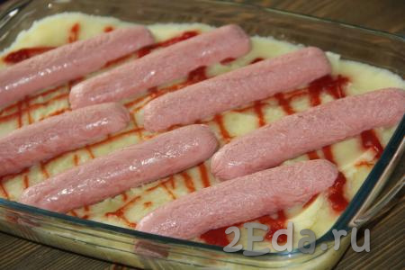 Нанести на картошку решётку (или полосочки) из кетчупа. Сверху выложить сосиски, предварительно очищенные от слюды (или оболочки). На поверхности каждой сосиски сделать несколько насечек острым ножом.