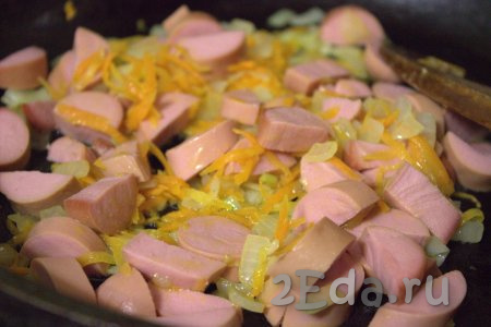 Очистить от плёнки, произвольно нарезать сосиски и добавить в сковороду к обжаренным овощам. Обжаривать сосиски с луком и морковью, помешивая, 5-7 минут.